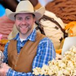 Smiling man in a cowboy hat holds a colorful Easter milkshake with bunny toppers, while popcorn and ice cream surround him.
