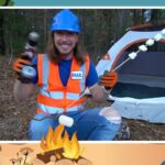 Man in a blue hard hat and orange safety vest sits by a camping tent in a forest, roasting marshmallows over a small fire.