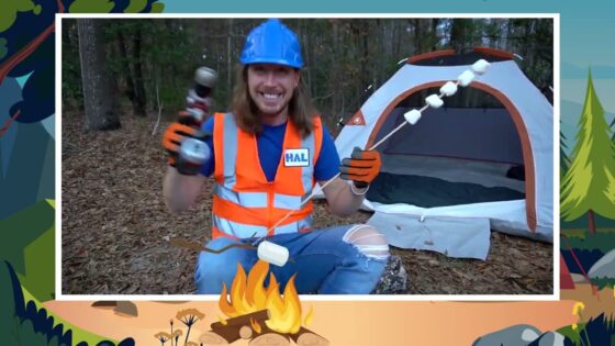 Man in a blue hard hat and orange safety vest sits by a camping tent in a forest, roasting marshmallows over a small fire.