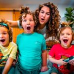 A woman with curly hair and three kids pose excitedly in a bright living room, making surprised faces; the child on the right holds a toy tool.