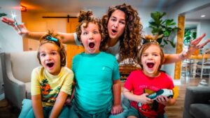A woman with curly hair and three kids pose excitedly in a bright living room, making surprised faces; the child on the right holds a toy tool.