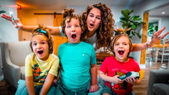 A woman with curly hair and three kids pose excitedly in a bright living room, making surprised faces; the child on the right holds a toy tool.