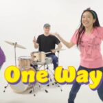 Band performance in a white studio: left guitarist, center drummer, right dancer in pink; large yellow 'One Way' text and a cartoon monkey with a guitar on the right.