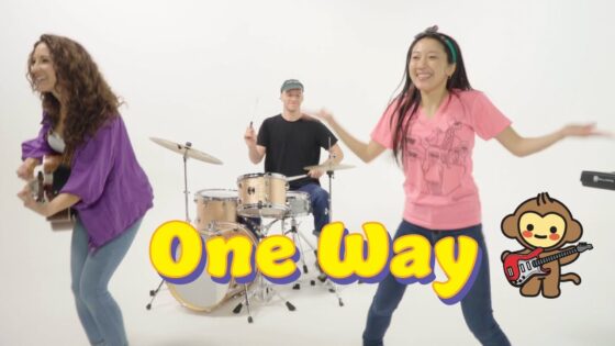 Band performance in a white studio: left guitarist, center drummer, right dancer in pink; large yellow 'One Way' text and a cartoon monkey with a guitar on the right.