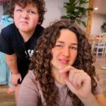 Woman with curly hair taking a selfie while a child behind her makes a funny face in a bright living room, wooden floor visible.