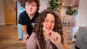 Woman with curly hair taking a selfie while a child behind her makes a funny face in a bright living room, wooden floor visible.