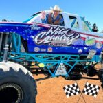 A blue monster truck with oversized tires and a smiling driver in a cowboy hat waves from the window on a dirt track, race flags at the front.