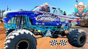 A blue monster truck with oversized tires and a smiling driver in a cowboy hat waves from the window on a dirt track, race flags at the front.
