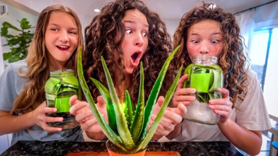 Three smiling girls pose with large glass jars of green liquid beside a potted plant on a kitchen counter.