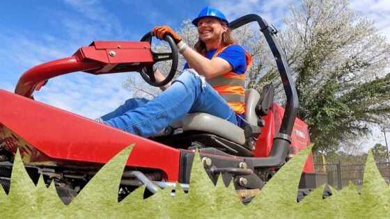 Smiling worker in a blue hard hat and orange safety vest driving a red ride-on lawn mower outdoors.
