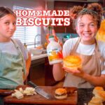 Two girls in aprons in a kitchen proudly display a homemade biscuit and a bottle of maple syrup by a cutting board with butter and a knife visible.