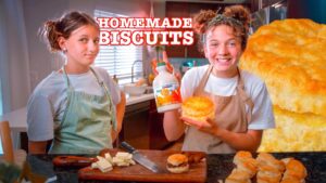 Two girls in aprons in a kitchen proudly display a homemade biscuit and a bottle of maple syrup by a cutting board with butter and a knife visible.