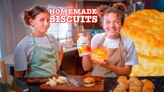 Two girls in aprons in a kitchen proudly display a homemade biscuit and a bottle of maple syrup by a cutting board with butter and a knife visible.
