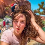 Woman takes a sunny outdoor selfie in a red rock desert, with a group of children playing in the background.