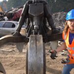 Construction worker in a blue hard hat and orange safety vest stands beside a giant hydraulic grapple in a scrapyard with wrecked cars in the background.