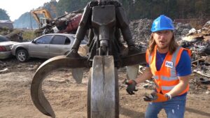 Construction worker in a blue hard hat and orange safety vest stands beside a giant hydraulic grapple in a scrapyard with wrecked cars in the background.
