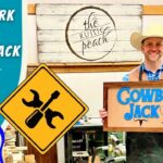 Smiling man in a cowboy hat holds a wooden 'Cowboy Jack' sign in a busy woodshop.