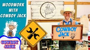 Smiling man in a cowboy hat holds a wooden 'Cowboy Jack' sign in a busy woodshop.
