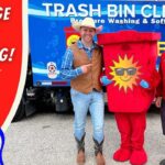 Promo photo for garbage can cleaning service with two men in western outfits posing with a red sun-logo trash can mascot in front of a blue Trash Bin Cleaning truck, promoting the service.