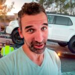 Man takes a selfie in front of a tow truck loading a white SUV onto a flatbed at sunset in a parking lot.