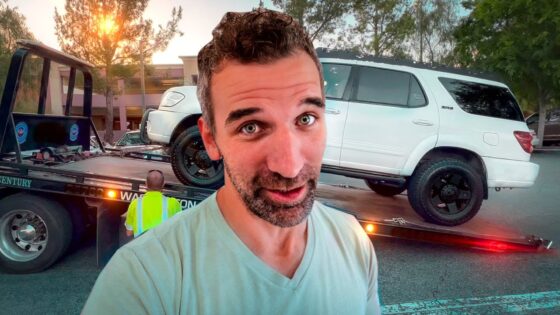 Man takes a selfie in front of a tow truck loading a white SUV onto a flatbed at sunset in a parking lot.