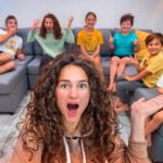 Woman with curly hair takes a surprised selfie in a living room as seven kids sit on a gray sectional behind her, cheering and posing for the camera.