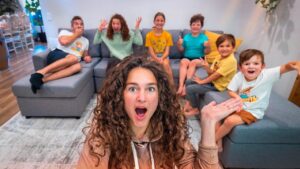 Woman with curly hair takes a surprised selfie in a living room as seven kids sit on a gray sectional behind her, cheering and posing for the camera.