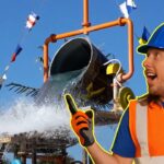 Construction worker in an orange safety vest and blue helmet points at a water splash from a slide or pipe structure behind him.