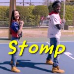 Two people stand back-to-back on a blue basketball court with a large yellow word 'Stomp' across the foreground, posing and smiling.