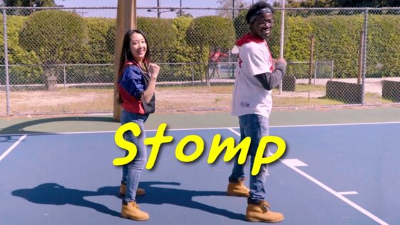 Two people stand back-to-back on a blue basketball court with a large yellow word 'Stomp' across the foreground, posing and smiling.
