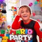 Smiling boy in red and black at a Mario Party birthday party with a large Mario figure, Bowser statue, balloons, and colorful toy bricks.