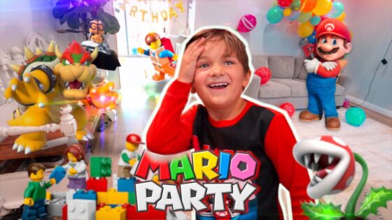 Smiling boy in red and black at a Mario Party birthday party with a large Mario figure, Bowser statue, balloons, and colorful toy bricks.
