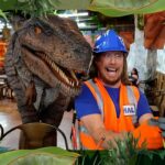 Man in blue hard hat and orange safety vest taking a selfie with a large dinosaur prop in a themed indoor setting.