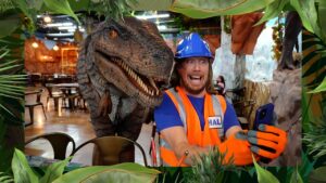 Man in blue hard hat and orange safety vest taking a selfie with a large dinosaur prop in a themed indoor setting.