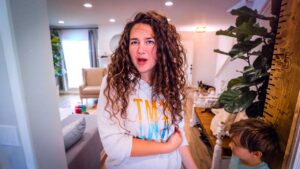 Mother with curly hair in a cozy living room, surprised expression, standing near a wooden growth ruler with a child nearby.