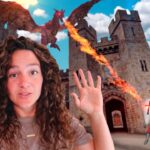 Woman with curly hair in the foreground at a castle as a red dragon breathes fire and a knight with a sword stands nearby.