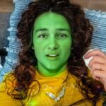 Person lying on a sofa with bright green face paint and a yellow shirt, holding a tissue.