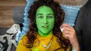 Person lying on a sofa with bright green face paint and a yellow shirt, holding a tissue.