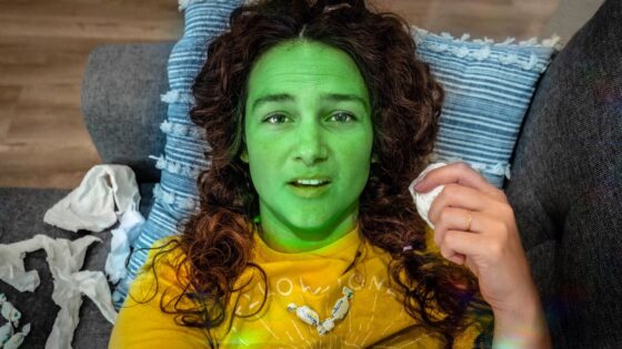 Person lying on a sofa with bright green face paint and a yellow shirt, holding a tissue.