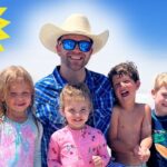 A smiling man in a cowboy hat and blue shirt with four kids on a sunny beach, waves in the background and a cartoon sun in the corner.