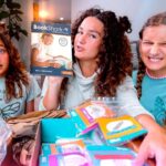 Three girls sit on a living room floor unboxing a BookShark homeschool kit, catalog visible.
