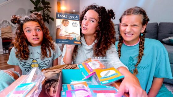Three girls sit on a living room floor unboxing a BookShark homeschool kit, catalog visible.