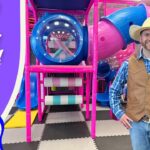 Man in a cowboy hat and vest posing in a colorful indoor playground, with a purple banner that reads 'Learn to Count to Ten!' on the left.