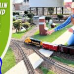 Miniature train layout with a smiling man in a cowboy hat kneeling beside tracks, pointing at the trains; left side shows bold green banner reading 'REAL TRAIN TOUR AND MODEL TRAINS' and a cartoon cowboy with 'COWBOY JACK' logo.