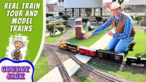 Miniature train layout with a smiling man in a cowboy hat kneeling beside tracks, pointing at the trains; left side shows bold green banner reading 'REAL TRAIN TOUR AND MODEL TRAINS' and a cartoon cowboy with 'COWBOY JACK' logo.