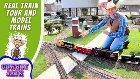 Miniature train layout with a smiling man in a cowboy hat kneeling beside tracks, pointing at the trains; left side shows bold green banner reading 'REAL TRAIN TOUR AND MODEL TRAINS' and a cartoon cowboy with 'COWBOY JACK' logo.