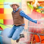Man in a cowboy hat and vest jumps beside a large toy shopping cart in a colorful playroom.3a8️