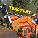 Orange compact tractor with front loader being operated outdoors by a man in a blue hard hat and safety vest; the word 'TRACTORS' is overlaid in yellow text.