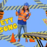 Two people in high-visibility orange safety gear, one at a keyboard and one in a suit, thumbs up with 'SAFETY SONG' on a blue background and construction tape pattern behind them.