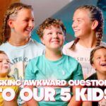 Five smiling kids posing indoors for a video thumbnail; bold title text across bottom reads 'Asking awkward questions to our 5 kids'.